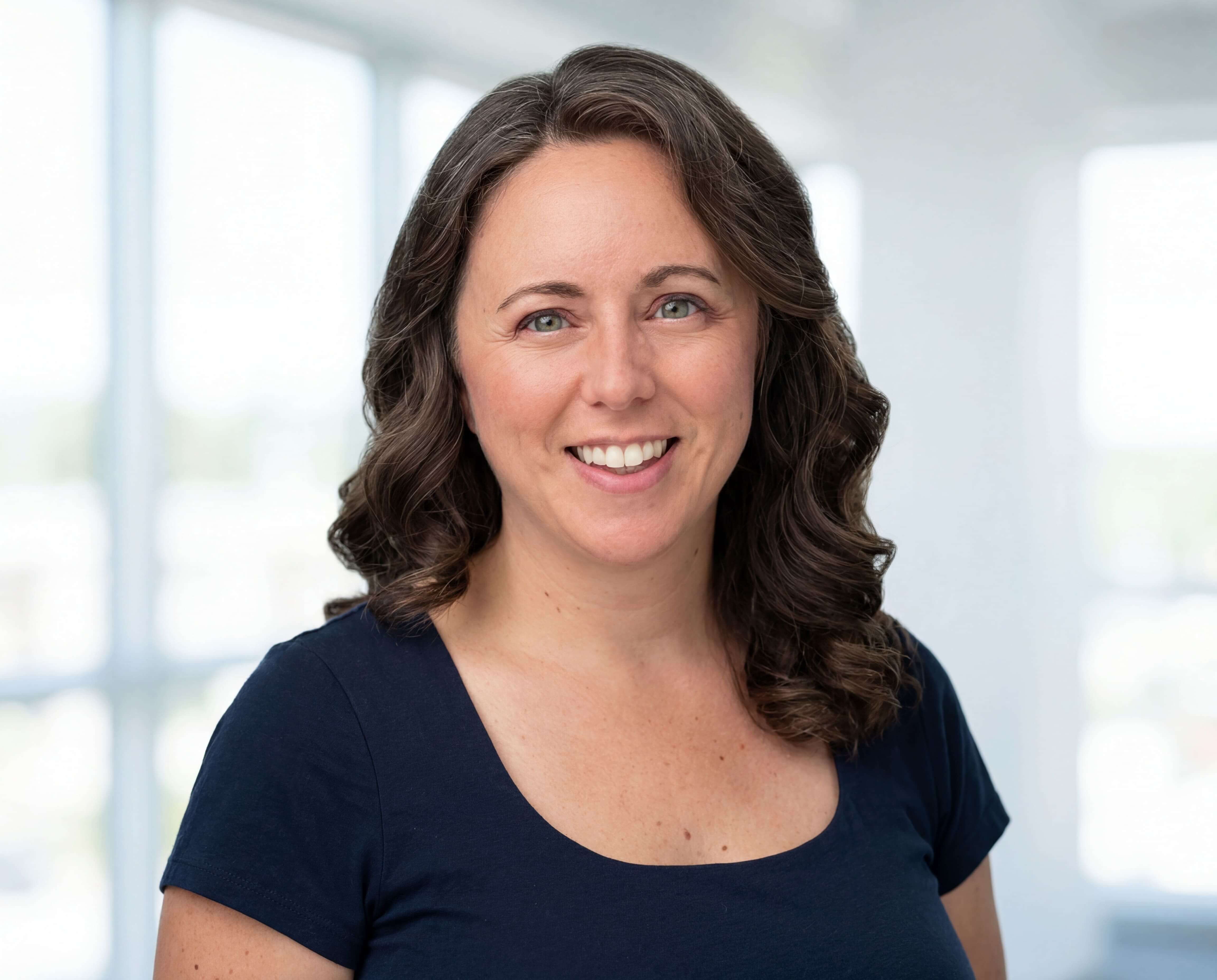 Chelsea Fournier, US trademark attorney and host of the Trademarks With Chelsea Podcast, in a navy blue short sleeve shirt standing in an office room with lots of windows behind her