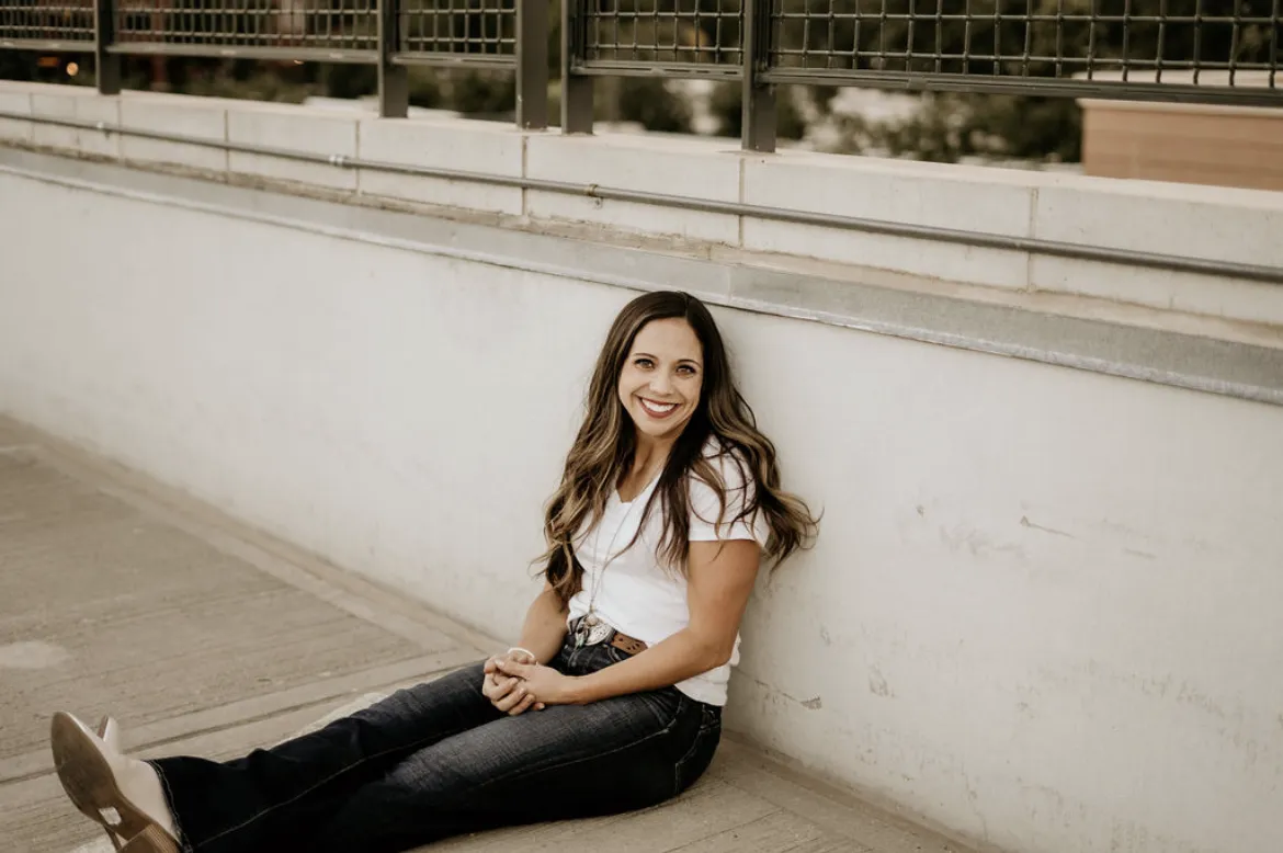 Picture of TinaB sitting on the ground with her legs screeched out leaning against a wall. Tina has long brown hair and is wearing a white  shirt with jeans. 