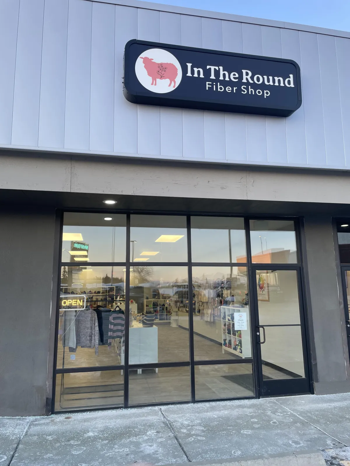 A charming storefront with large windows, a hand-painted sign reading 'In the Round Fiber Shop,' and planters filled with seasonal flowers. The shop is set against a backdrop of Anchorage’s cityscape and distant mountains.