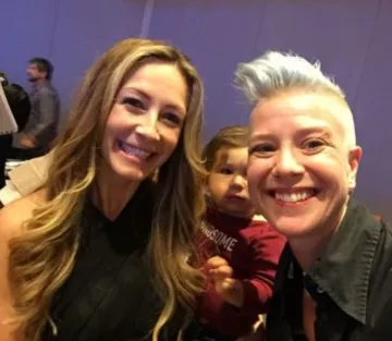 Brigitta Hoeferle and Shanda Sumpter smile for a photo with a child in the background at a lively event gathering.