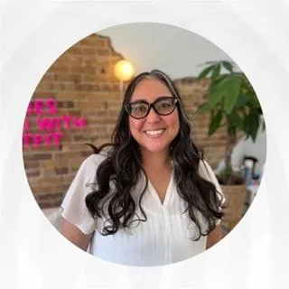 A smiling woman with long dark wavy hair and black glasses, wearing a white blouse, in a circular frame. Behind her is an exposed brick wall with pink neon signage, warm lighting, and a green plant. Headshot of Jennifer Anne Garcia, Career Strategy Coach at bestresumecoach.com. #BestResumeCoach #CareerStorytelling
