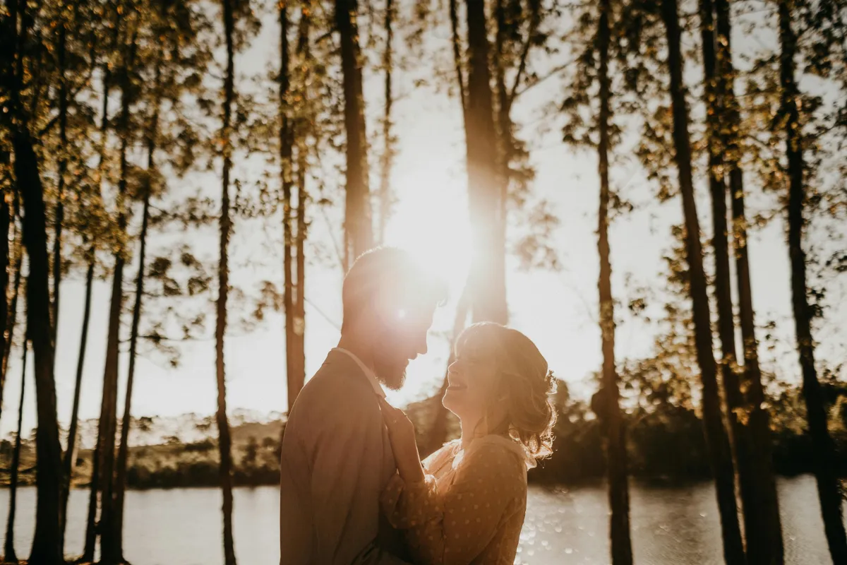 Smiling couple in the woods by a lake, gazing into each other’s eyes.