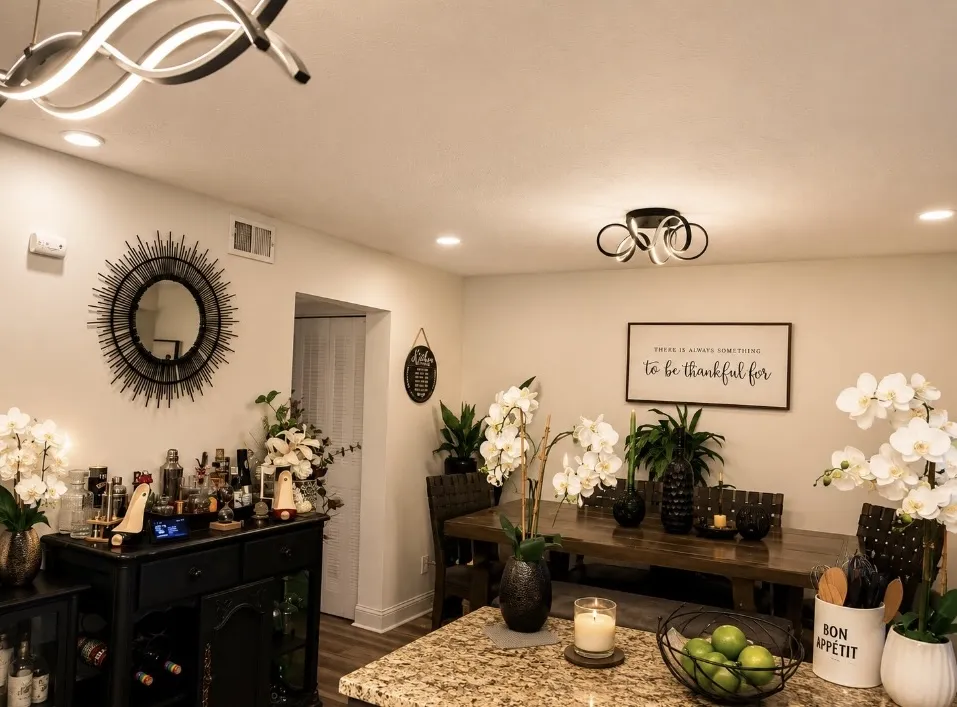 Warm dining and entertaining space with wood dining table, black furnishings, white orchids, greenery, bar area, and modern lighting.