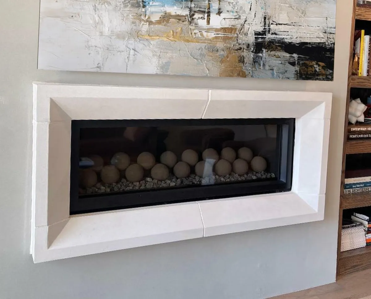 The Picture Frame, a modern linear fireplace mounted on a wall like a piece of art, with a beveled stone frame and ceramic fire ball display.