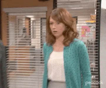 Woman in a teal cardigan stands in an office, looking surprised as she turns toward someone off-camera.