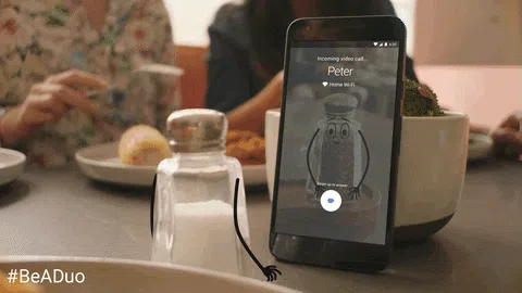 A smartphone on a dining table displays an incoming video call from Peter while people eat in the blurred background.
