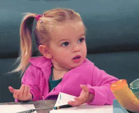 Young girl in a bright pink jacket sitting at a table with her hands raised, looking surprised and confused.