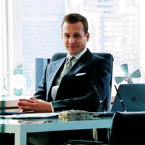 Man in a dark suit sits confidently behind a modern office desk, looking toward the camera with his hands clasped.