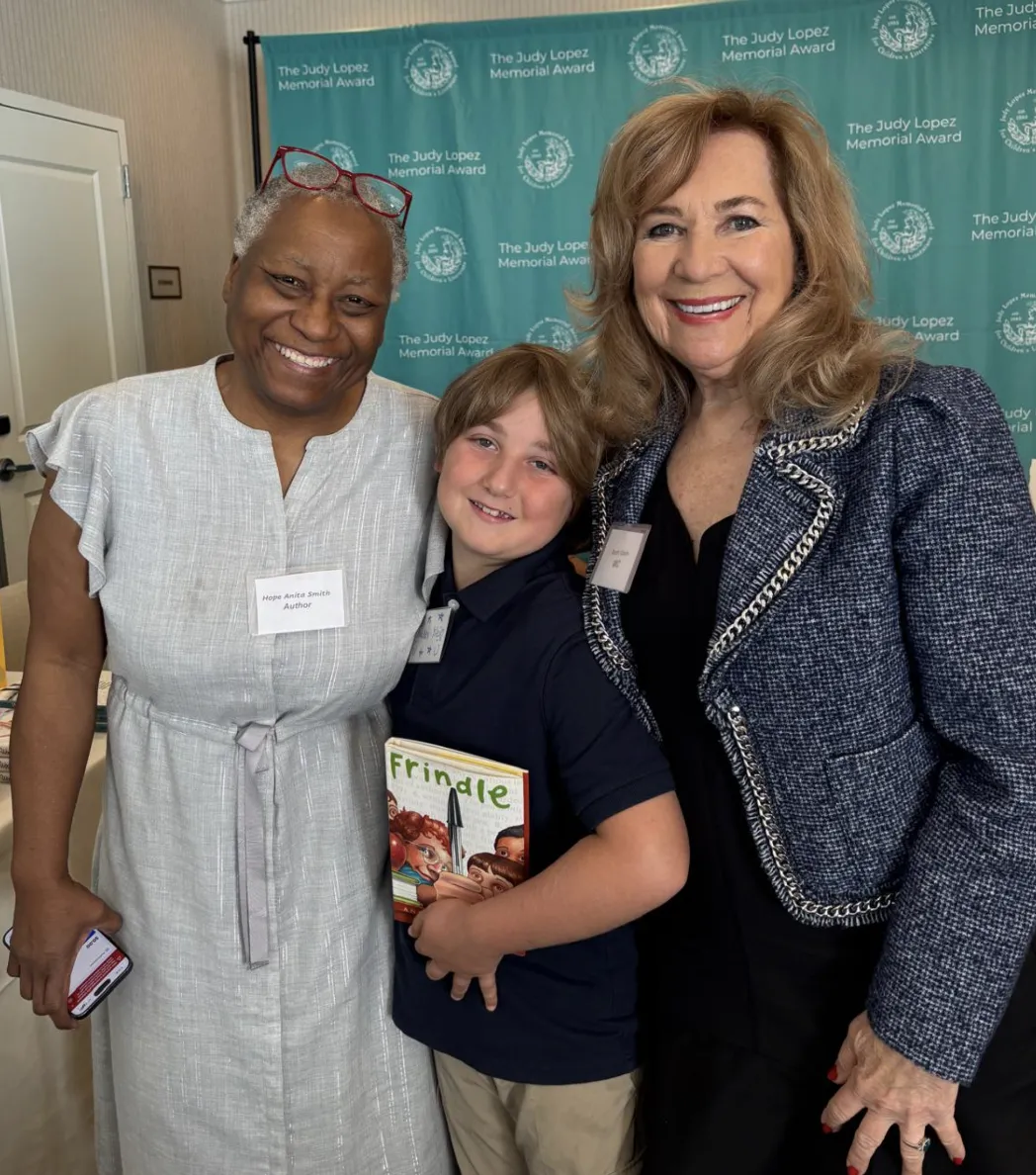 Hope Anita Smith, a young boy holding a book, and Ruth Klein smiling together in front of the Judy Lopez Memorial Award backdrop.