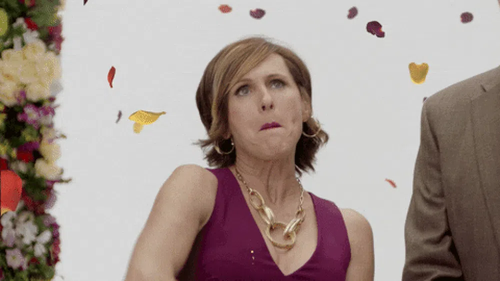Woman in a sleeveless purple dress making a playful, exaggerated facial expression as colorful flower petals fall around her against a white background.