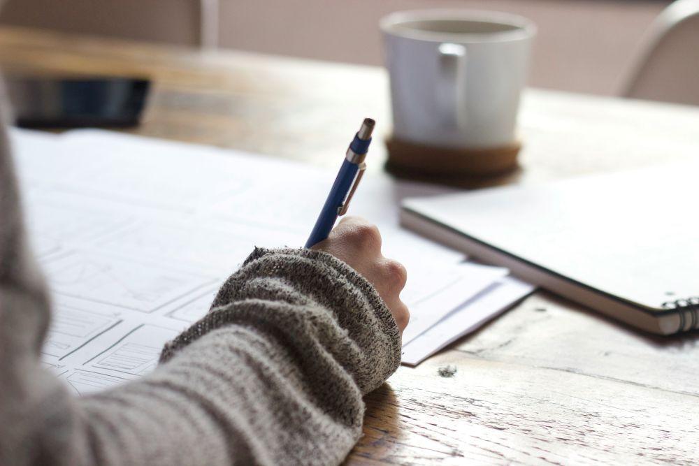 Person writing with a blue pen on printed papers at a wooden table, with a coffee mug and closed notebook nearby.