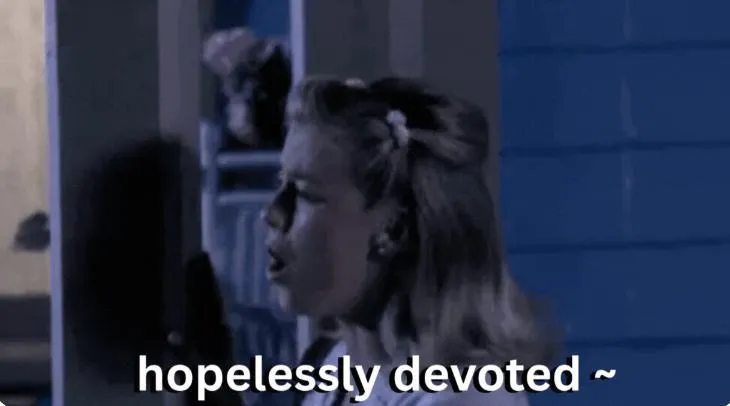 Woman with curled blonde hair sings in a dimly lit scene as the words “hopelessly devoted” appear across the bottom.