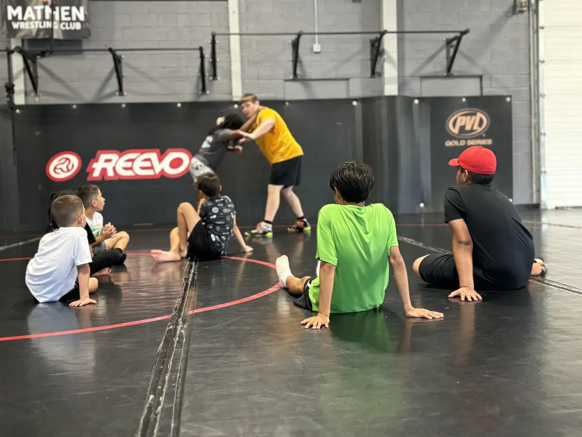 A smiling child wearing a Battle Arts Academy camp t-shirt and holding a lunch tray, with art supplies and other campers in the background.