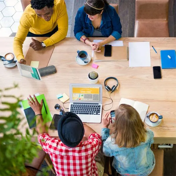 Overhead view of a diverse group of people collaborating around a desk with laptops, coffee, and notebooks, symbolizing teamwork in crafting marketing success stories. The image complements content about creating innovative, client-centered marketing strategies that help businesses stand out and leave a lasting impact.