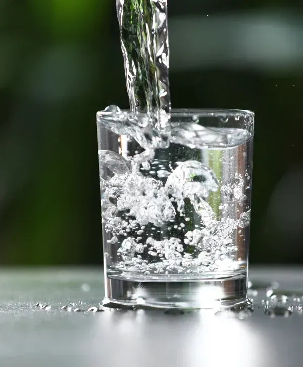 A stream of clear water pouring into a clear glass, creating many bubbles and splashing onto a dark surface.