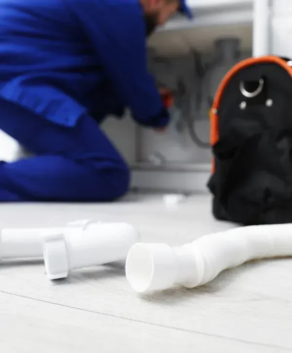 A blurry image of a plumber in a blue uniform working under a sink, with new white pipes in the foreground.