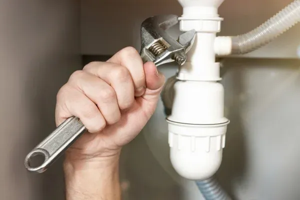 A hand wearing a yellow glove adjusts a gray P-trap pipe under a sink, correcting plumbing with visible joints.