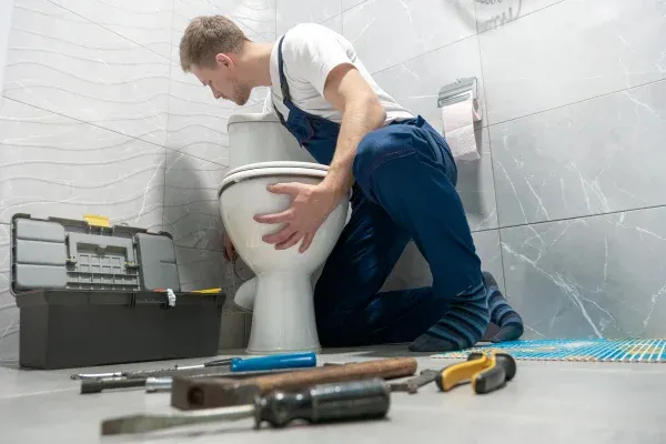A male plumber in blue overalls inspects and repairs a toilet in a home bathroom with tools scattered around.