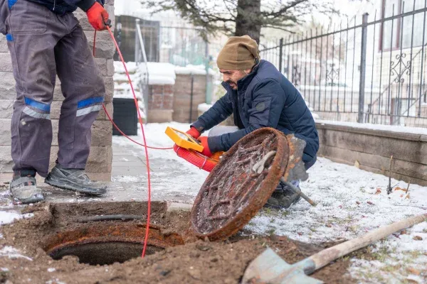 Two utility workers inspecting a manhole with specialized equipment in a snowy, residential outdoor setting.