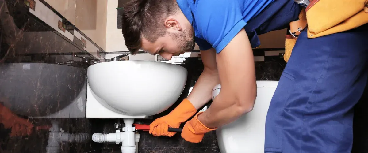 A male plumber in blue uniform and orange gloves fixing a white bathroom sink with a wrench.