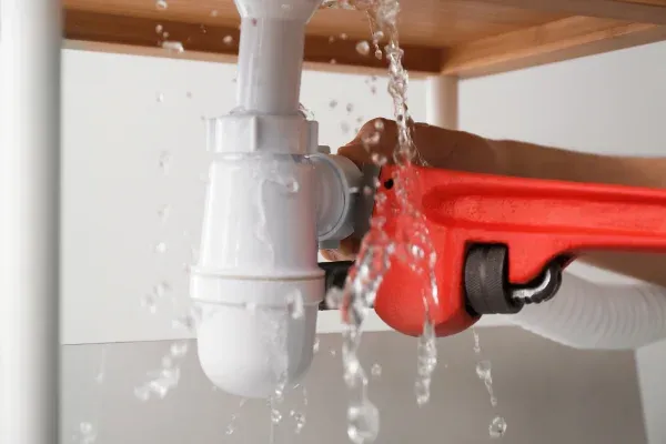 A hand using a red pipe wrench to tighten a leaking white P-trap under a wooden shelf with water splashing.