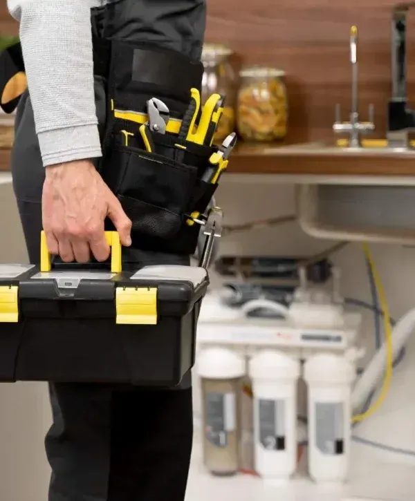 A cropped shot of a plumber carrying a toolbox and wearing a tool belt, standing in a kitchen.