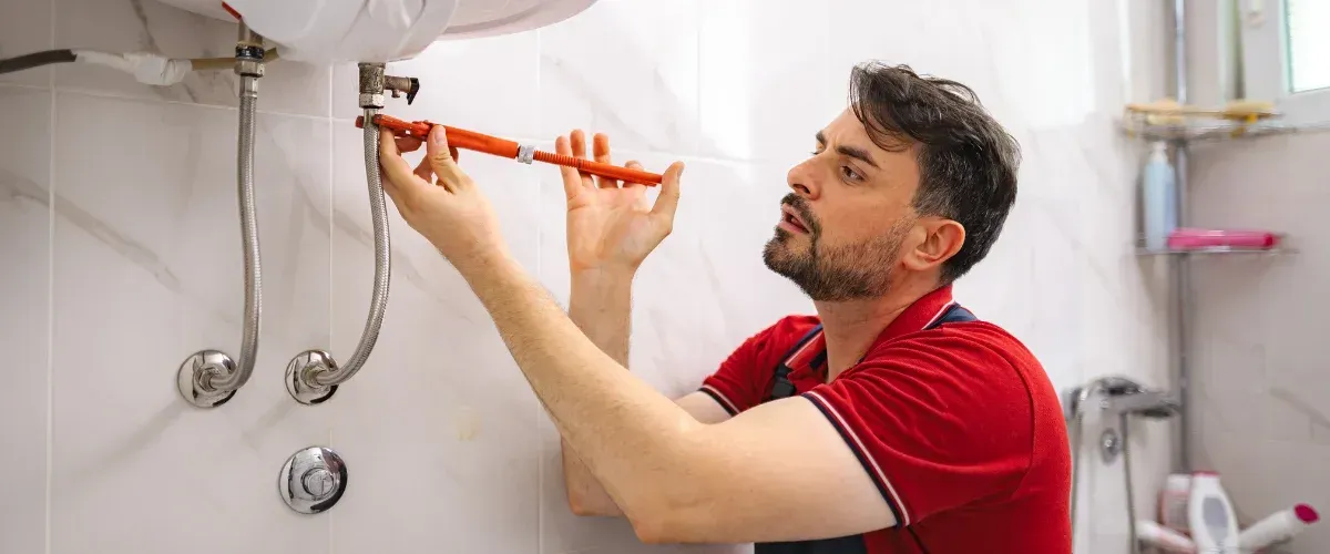 A male plumber with a beard wearing a blue shirt and grey overalls, using a wrench to repair a sink pipe.