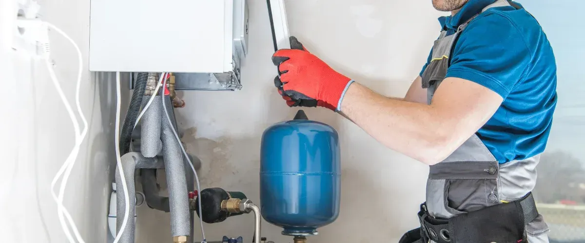 A gloved plumber in blue and grey work clothes installing or maintaining a white boiler and blue expansion tank.