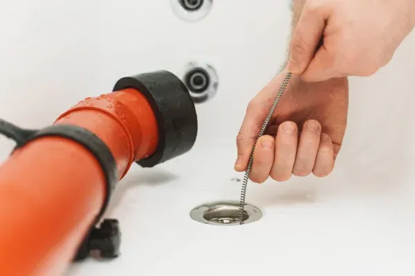 A close-up of a person's hands using a drain snake to clear a clog in a white bathtub drain.