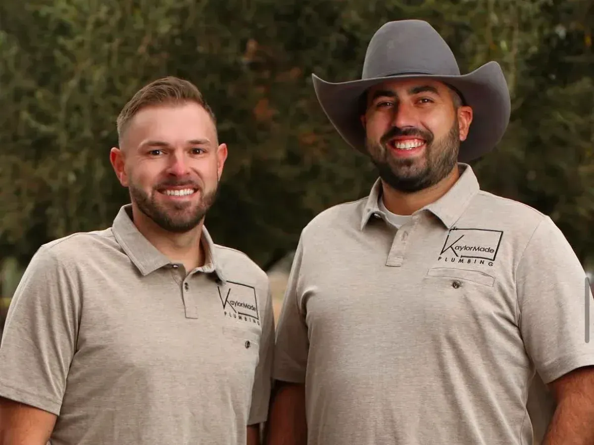 Two smiling men in KaylorMade Plumbing polo shirts, one wearing a gray cowboy hat, stand outdoors.