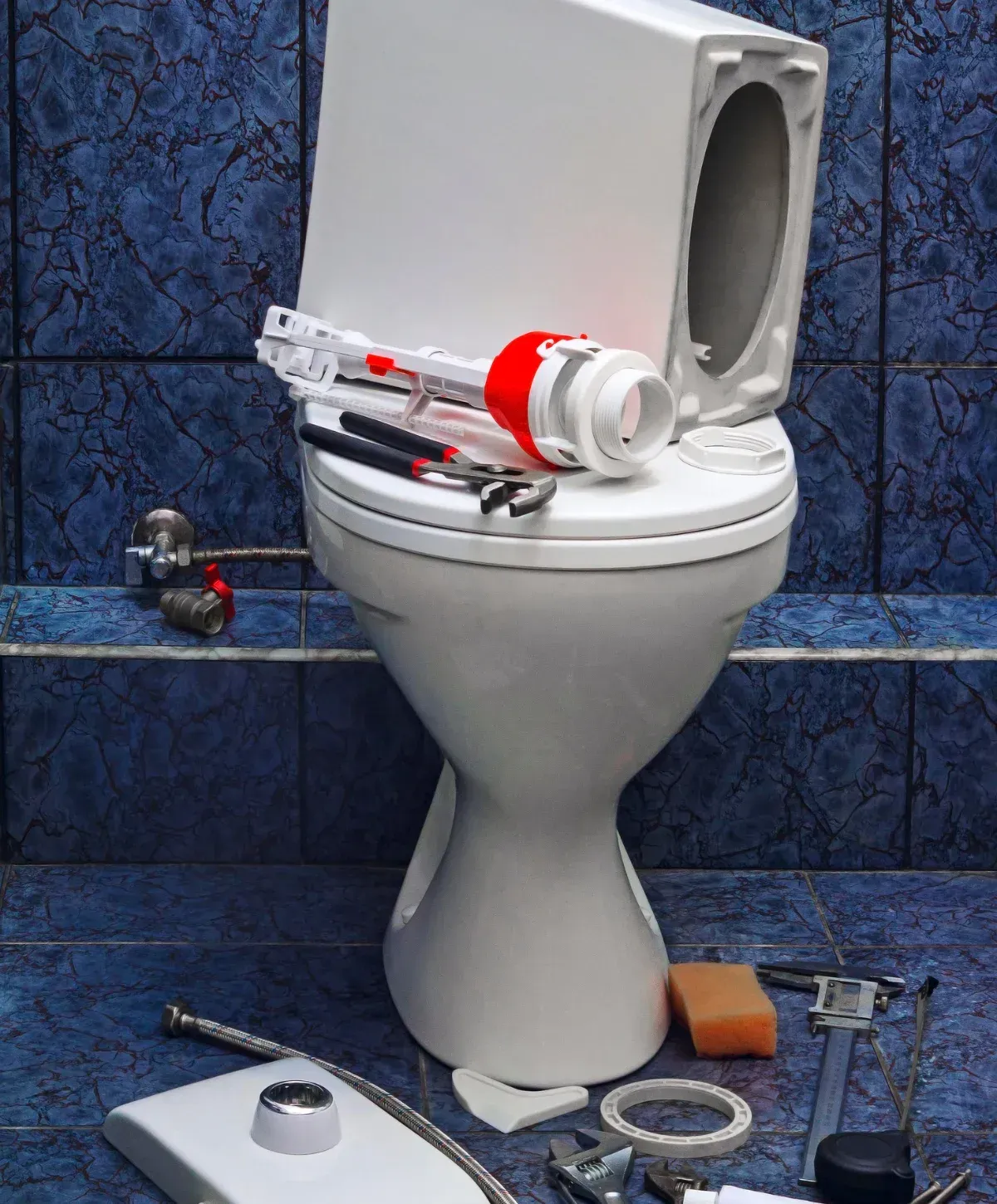 A disassembled white toilet with plumbing parts, tools, and an orange sponge scattered on blue tiled floor and shelf.