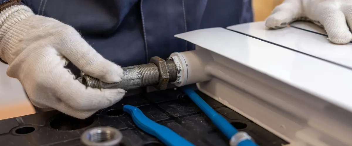 A close-up of gloved hands installing a threaded metal pipe into a white radiator or heating unit, with blue tools nearby.
