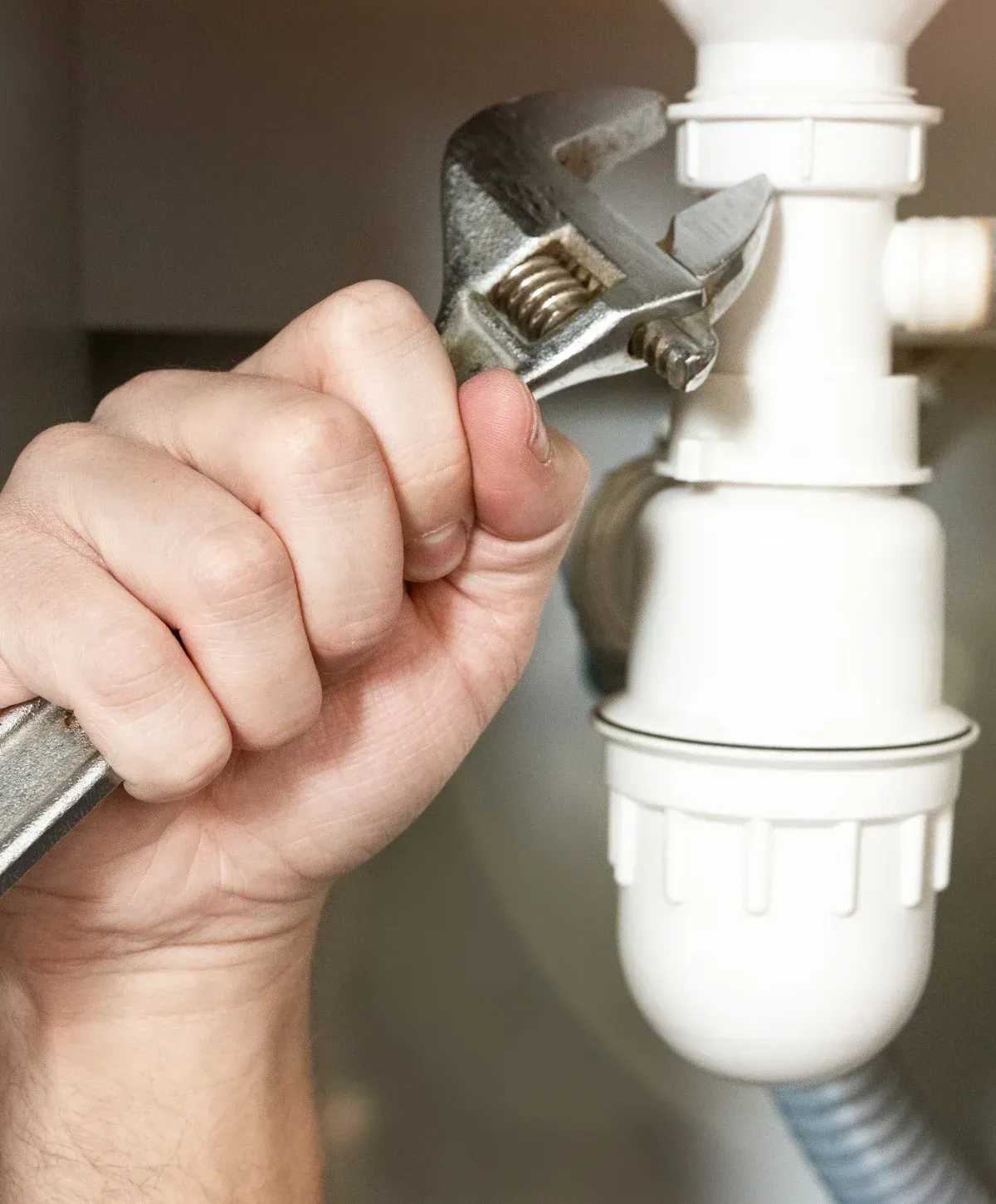 A person's hand uses an adjustable wrench to tighten a white plastic sink trap pipe, likely for plumbing repair.