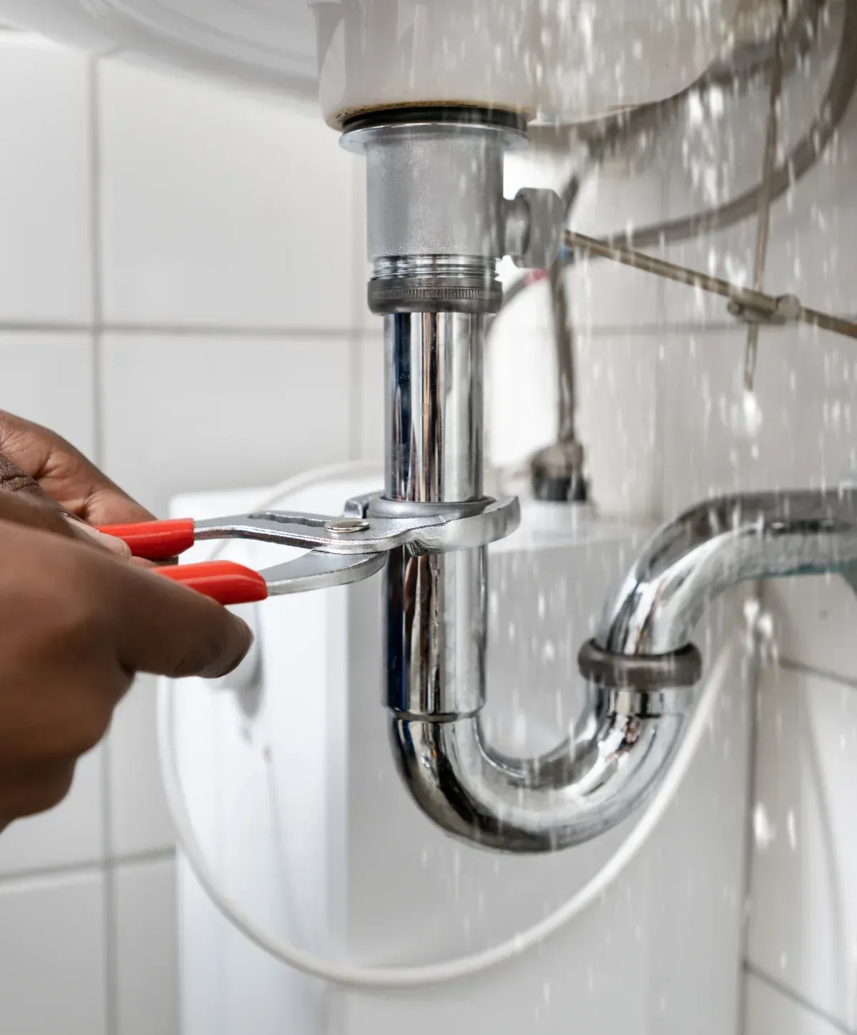 A person's hands using a red-handled wrench to fix a leaking chrome sink drain pipe with water splashing.