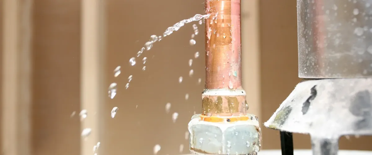 A close-up of a copper pipe with water spraying forcefully from a leak, showing individual drops.