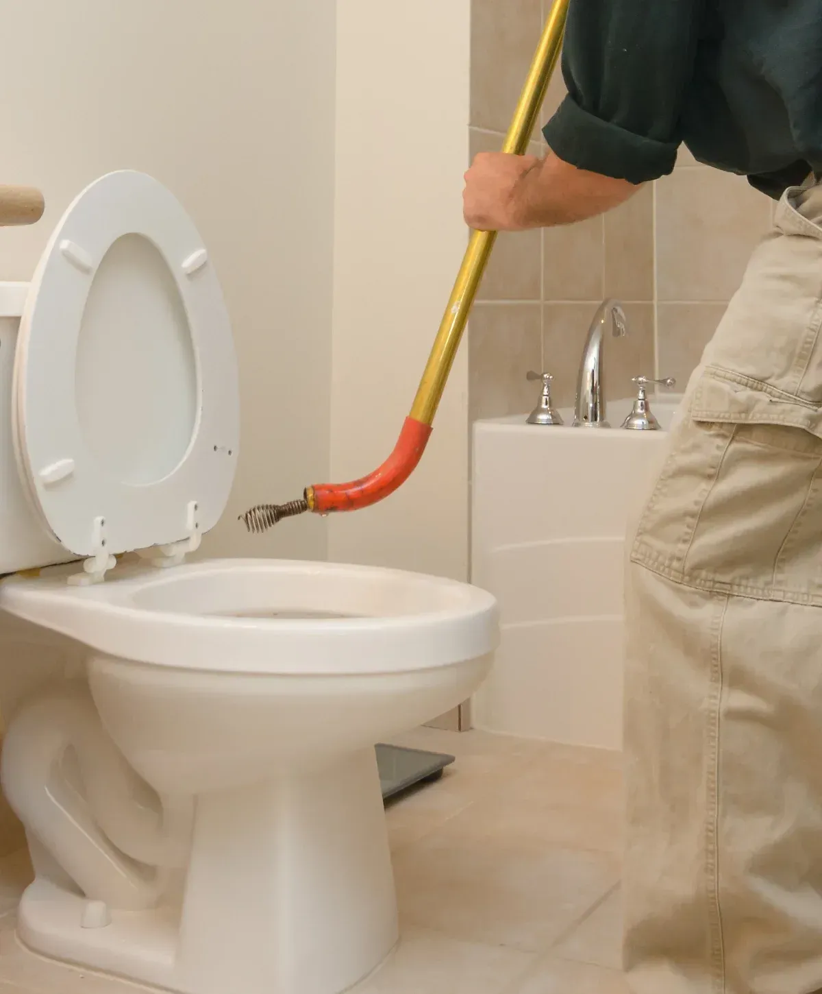 A person in khaki pants and a dark shirt using a toilet auger to clear a clogged toilet in a bathroom.