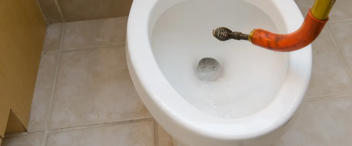 A close-up, high-angle view of an orange plumbing snake inserted into a white toilet bowl with swirling water.