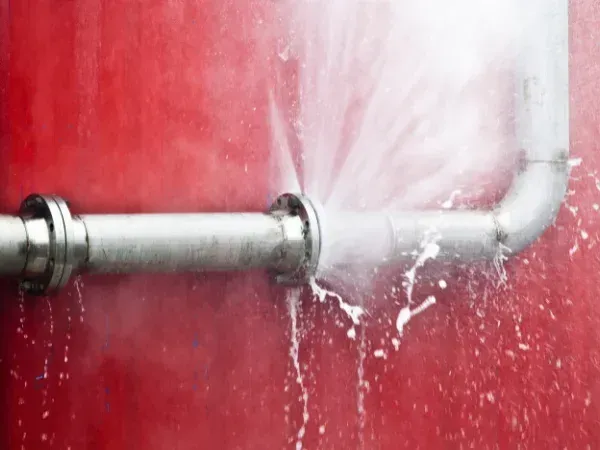 Water forcefully gushes from a burst metal pipe connection, creating a white spray against a vibrant red background.