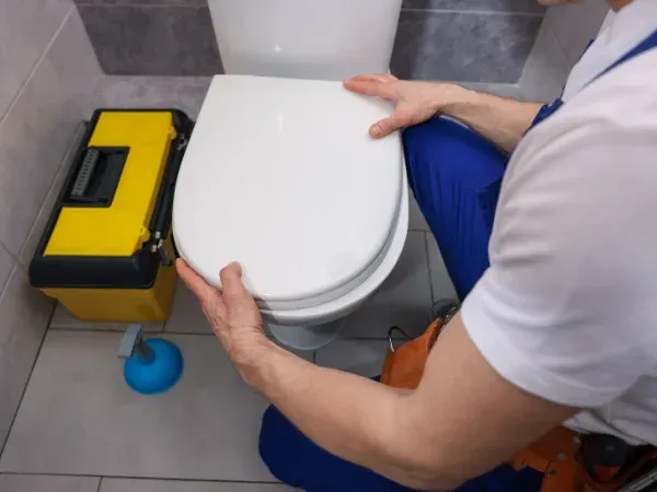 A close-up shot of a person in blue work pants installing or adjusting a white toilet seat in a bathroom.