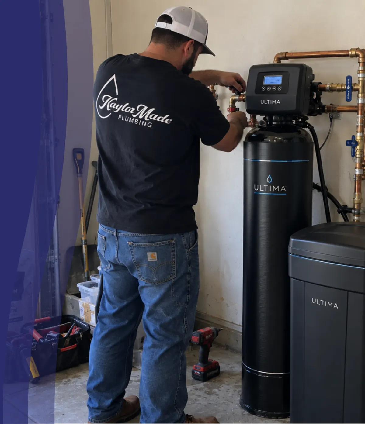 A plumber wearing a black 'Kaylor Made Plumbing' shirt works on a black water softener system with copper pipes.