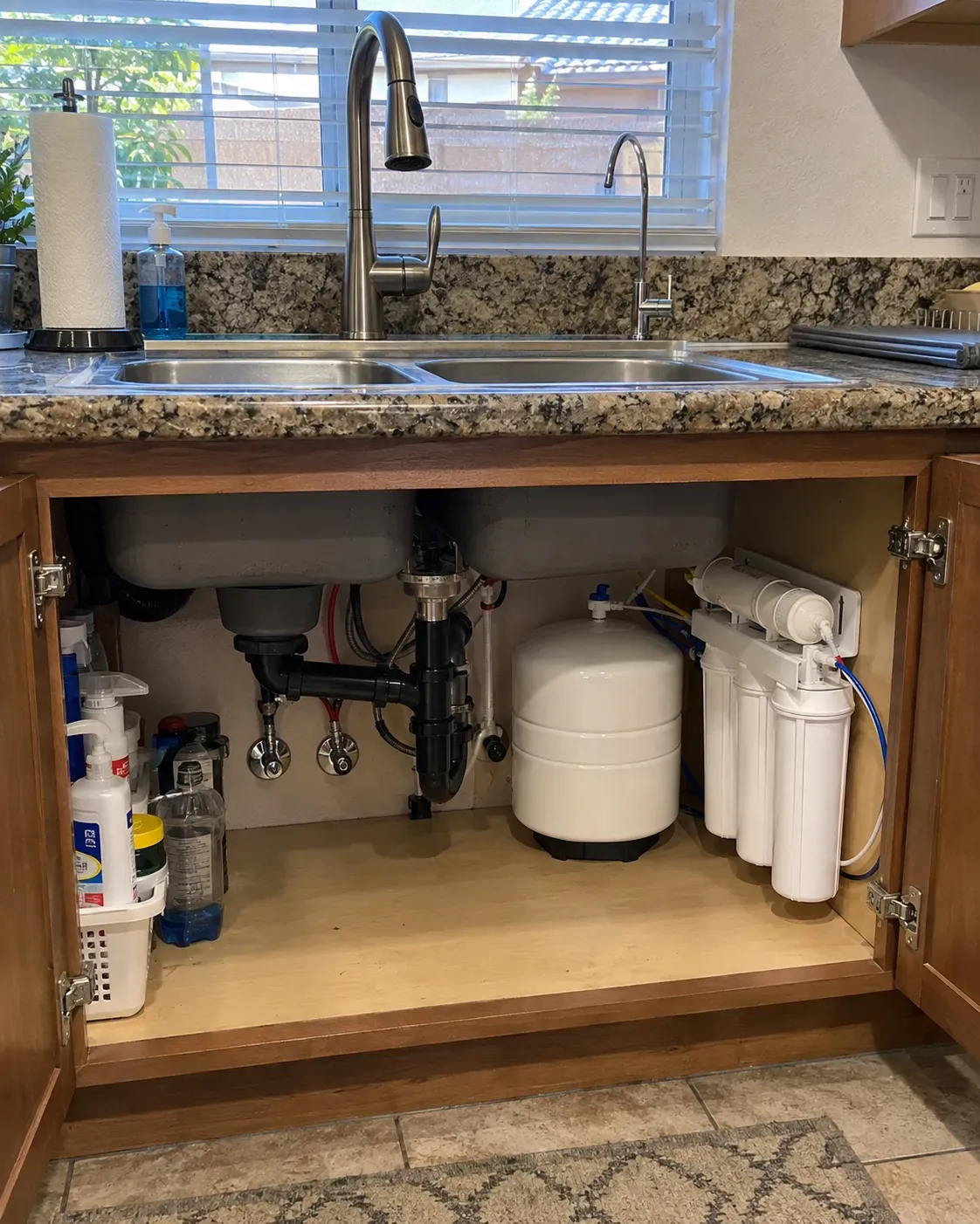 An open under-sink cabinet in a kitchen reveals a white reverse osmosis water filtration system and pipes.