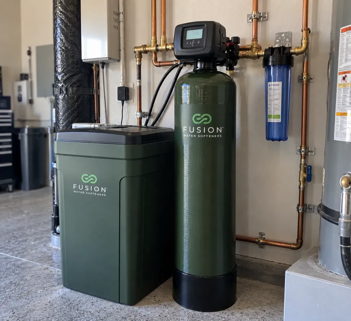 A Fusion Water Softeners system with a dark green brine tank and tall softener tank, alongside a blue Pentair filter, installed with copper pipes in a utility room.