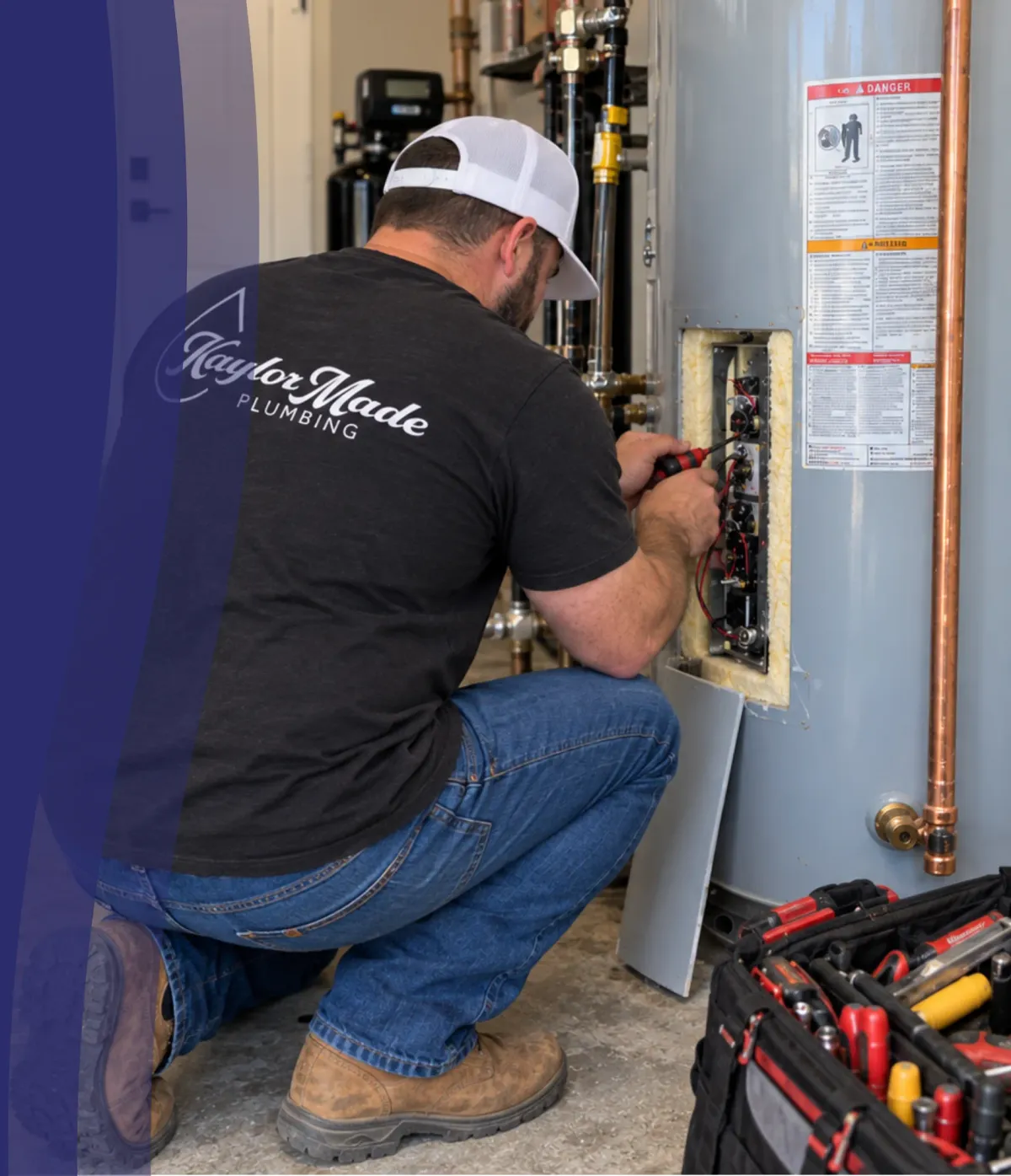 A plumber in a white cap and black shirt kneels to repair the electrical panel of a water heater.