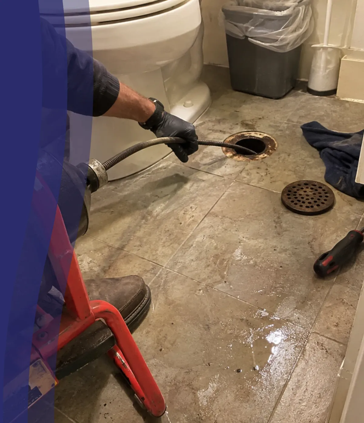 A gloved plumber uses a drain snake to clear a clog from a floor drain next to a toilet in a wet bathroom.