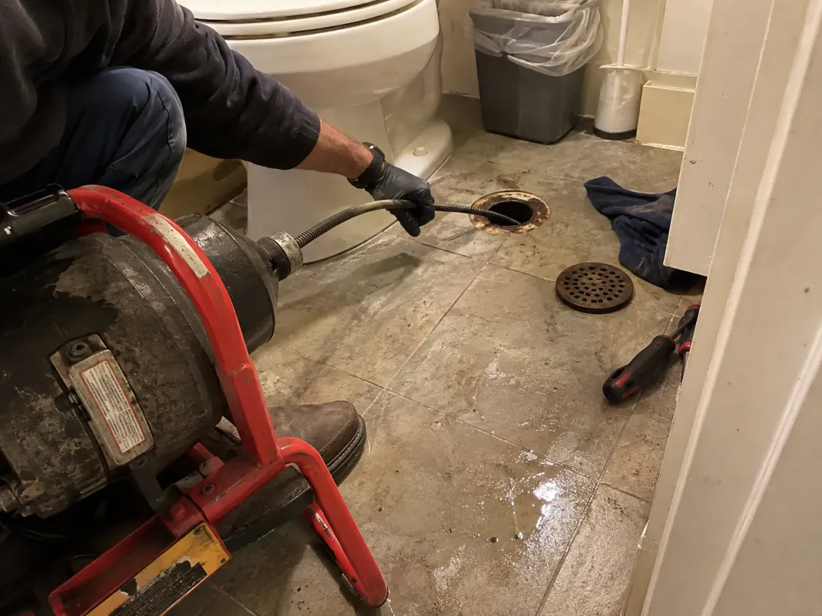 A gloved plumber uses a heavy-duty drain auger to clear a floor drain in a tiled bathroom, with a toilet nearby.
