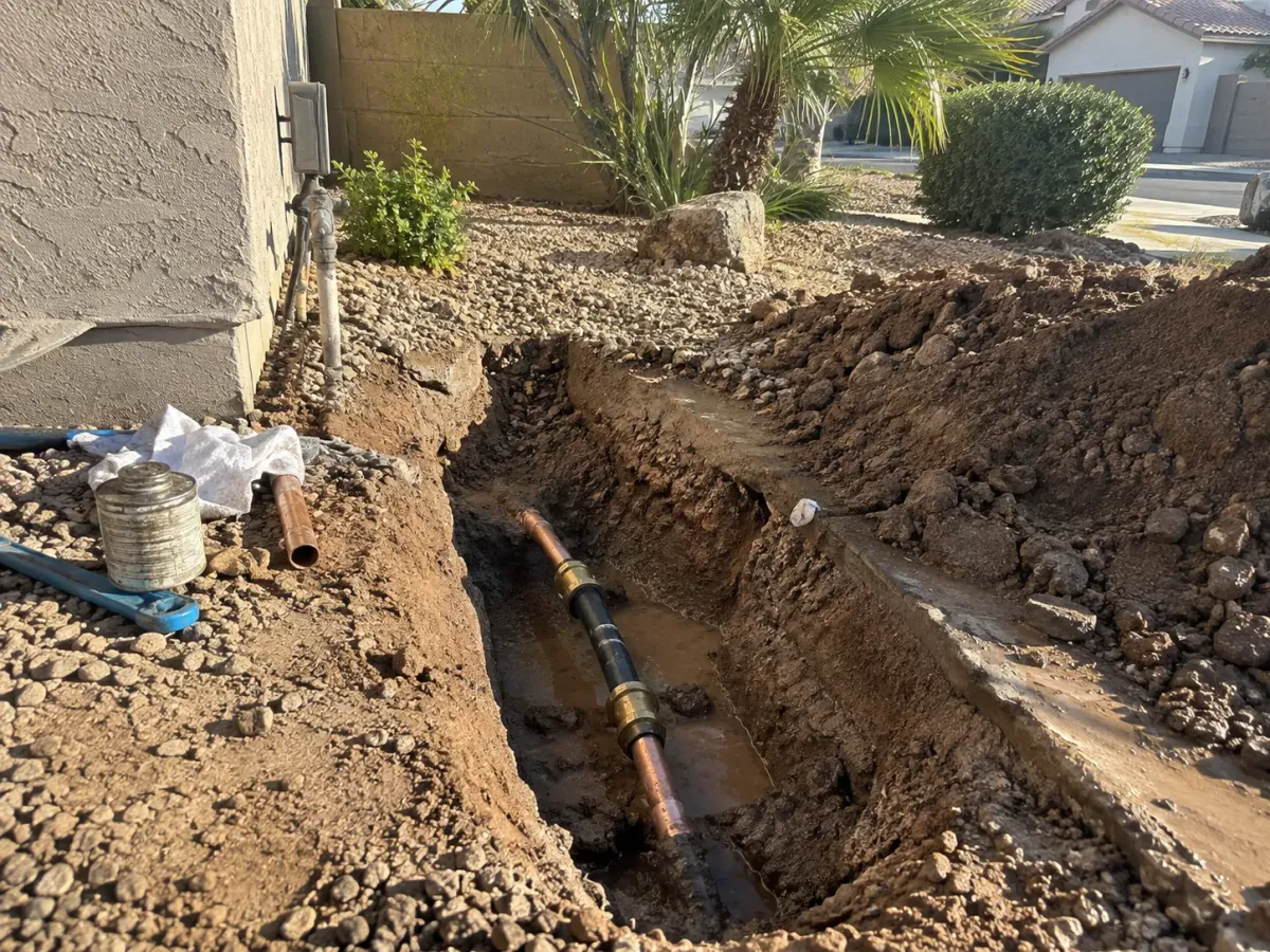 A trench dug next to a house foundation shows a repaired water pipe with a copper segment and muddy water.