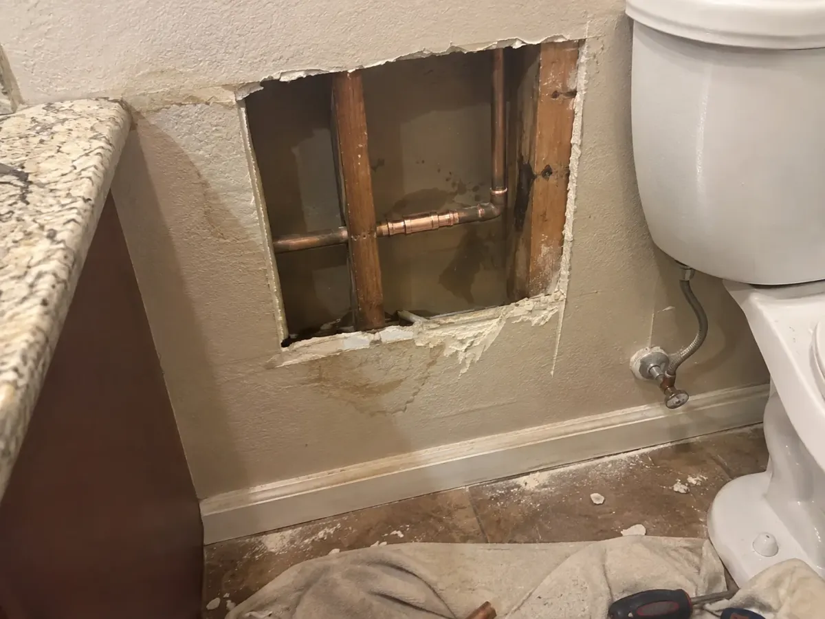 A drywall access hole reveals copper pipes and wooden studs behind a wall, next to a toilet and granite countertop.