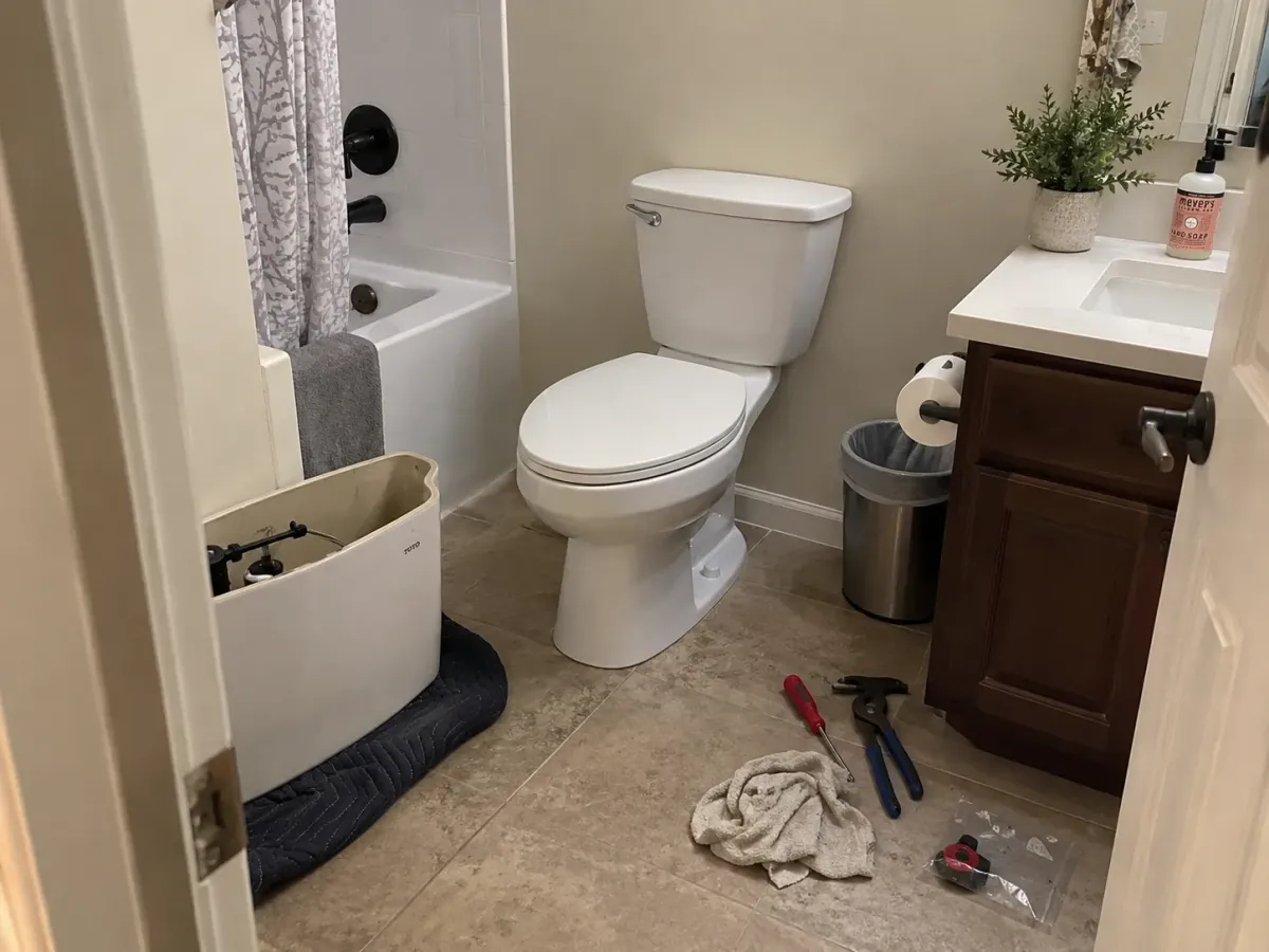 A bathroom scene showing a white toilet, its tank detached on the floor, and repair tools nearby.