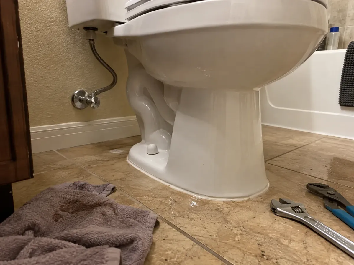A leaky white toilet with a used towel and plumbing tools (wrench, pliers) on the tiled bathroom floor, suggesting a repair.