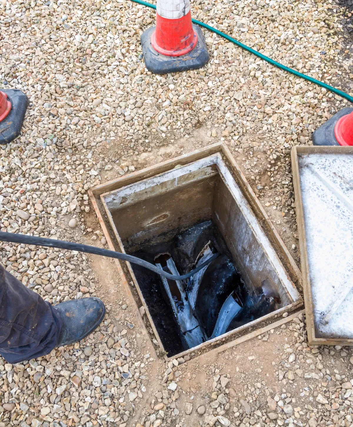 A person's foot near an open manhole on gravel, with a hose entering the dark, water-filled inspection chamber.